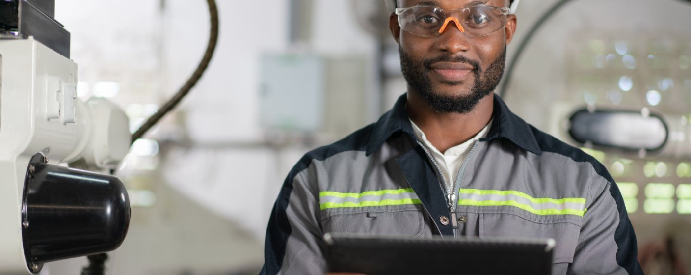 Colaborador segurando tablet vestido com 1 roupa cinza e azul, 1 listra na horizontal amarela, capacete epi branco e óculos de proteção