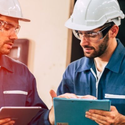 2 funcionários com jaqueta azul, capacete epi branco, óculos de proteção manuseando aparelho retangular 1 na cor cinza e outro azul