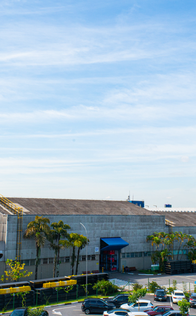 Galpão industrial com estrutura robusta cinza, toldo azul na entrada, a sua frente possui estacionamento com carros e arvores a sua volta