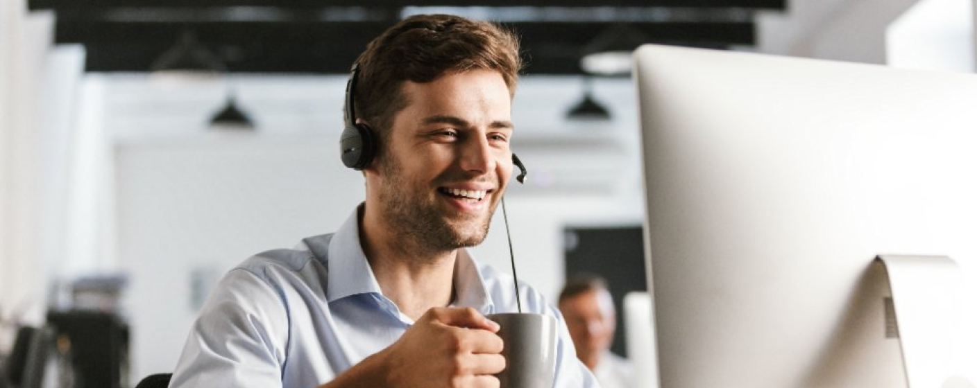 Homem prestando suporte técnico remoto, vestindo camisa branca, fone de ouvido na cabeça, xicara sobre a mão em frente ao computador