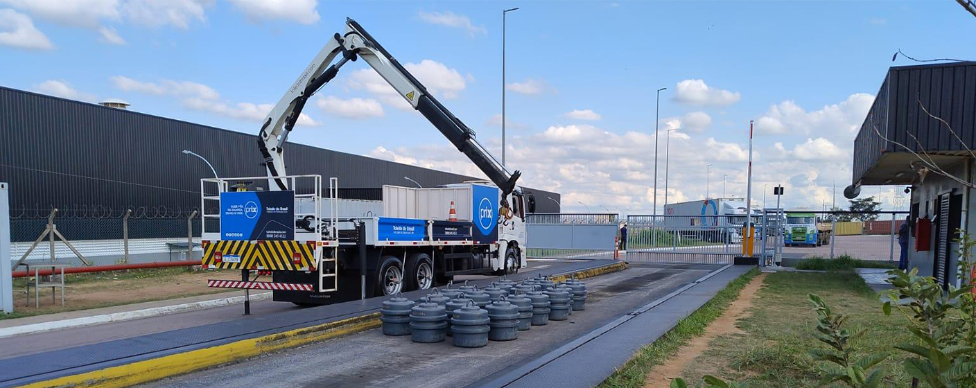 Caminhão UMC (Unidade Móvel de Calibração) estacionado sobre uma balança rodoviária instalada em uma empresa. No fundo, a paisagem conta com plantio de grãos e céu azulado.