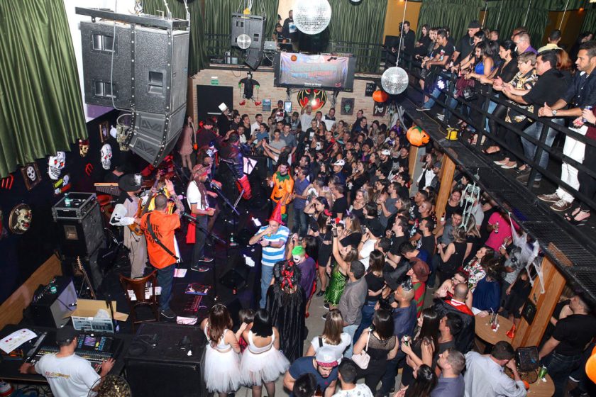 Colaboradores da Toledo do Brasil fantasiados aproveitam apresentação musical ao vivo em evento corporativo temático com decoração de Halloween em ambiente interno lotado.