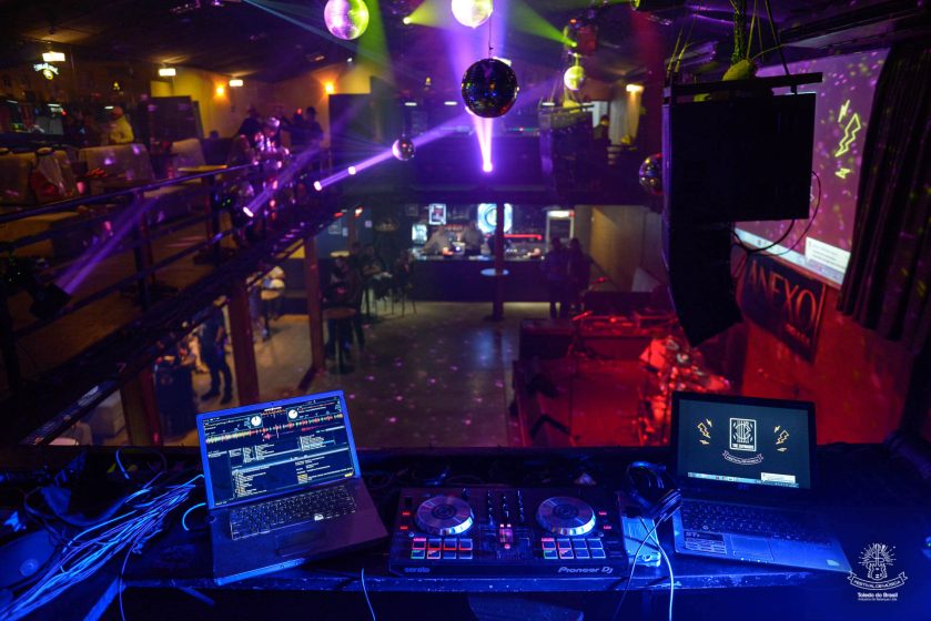 Vista da cabine do DJ com equipamentos e notebooks iluminados por luzes roxas durante evento noturno da Toledo do Brasil com pista de dança decorada e palco preparado para apresentações.