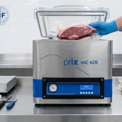 Seladora a vácuo Prix Vac 42B e balança industrial Toledo do Brasil sobre bancada de aço inox em uma cozinha profissional, com operador embalando carne a vácuo.
