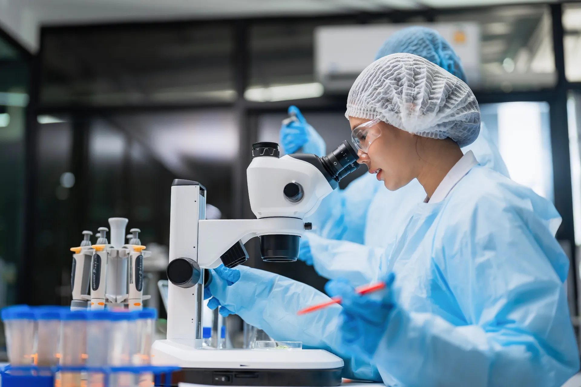 Profissional com touca, óculos de proteção e luvas utilizando microscópio em laboratório.